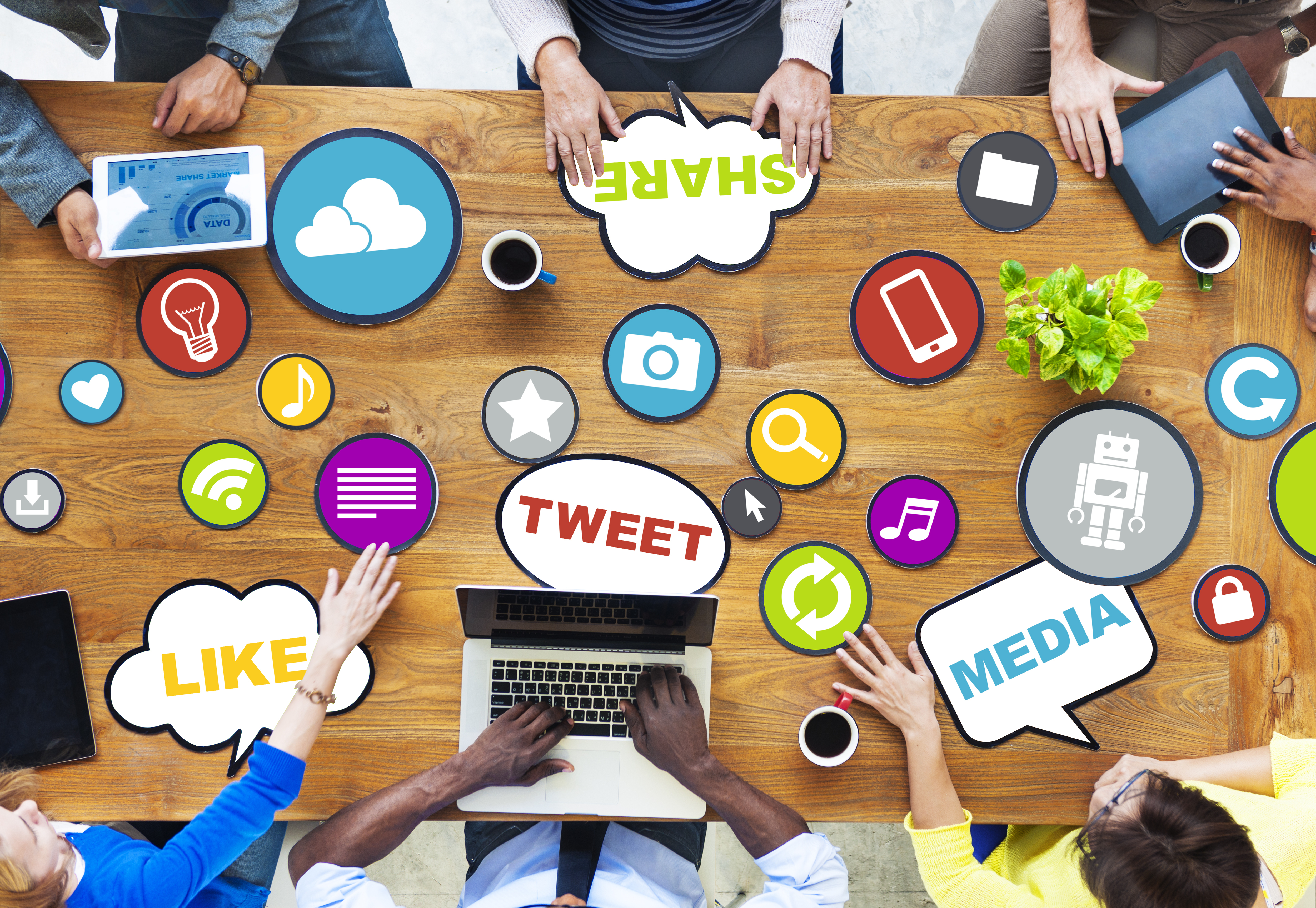 Top-down view of a tabletop with icons related to social media on the surface, and people sitting around the table with laptops and tablets.